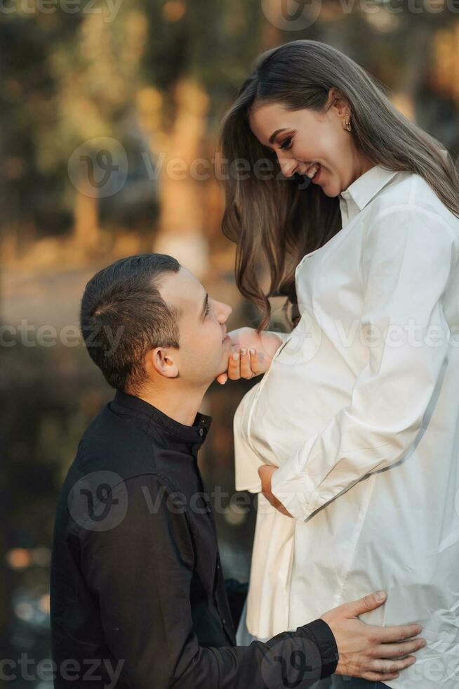 jong zwanger vrouw in een wit shirt. de man zetten zijn handen Aan zijn vrouwen maag en leunde tegen zijn oor. foto