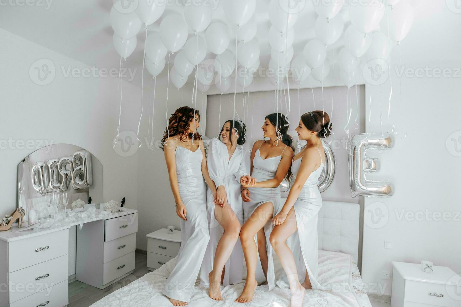 jong bruidsmeisjes in wit zijde jurken Aan een bed in de bruid kamer. mooi Dames vieren vrijgezellin partij zittend Aan bed. foto