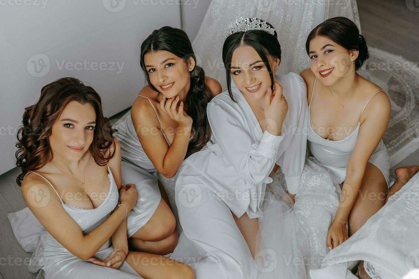 jong bruidsmeisjes in wit zijde jurken Aan een bed in de bruid kamer. mooi Dames vieren vrijgezellin partij zittend Aan bed. foto