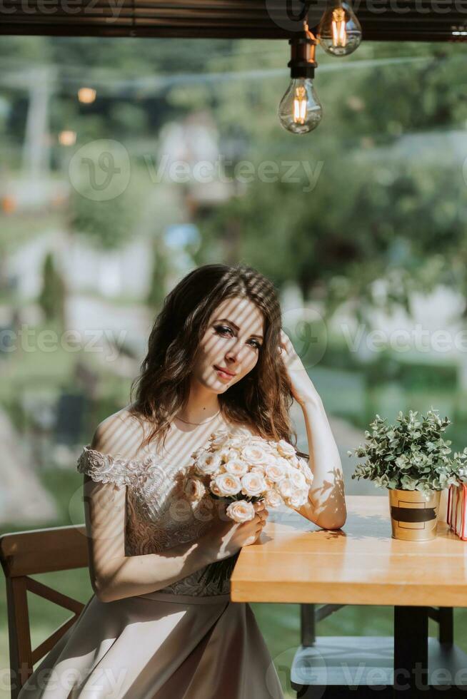 een jong elegant vrouw in een beige jurk met een boeket van bloemen in haar handen is zittend in een mooi licht in een cafe in Spanje foto