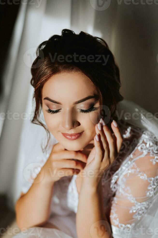 de ochtend- van de bruid in een wit gewaad en een elegant kapsel. een volwassen meisje zit Aan een stoel en vastmaakt een oorbel, op zoek in de camera lens en glimlachen foto