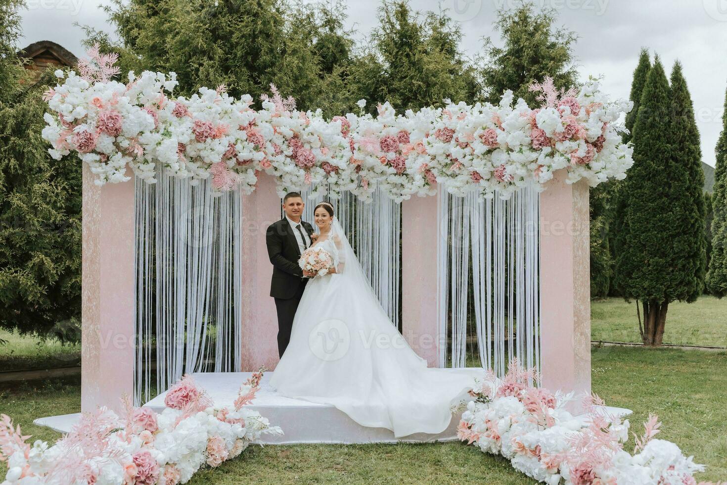 aantrekkelijk bruid en bruidegom Bij de ceremonie Aan hun bruiloft dag met een boog gemaakt van roze en wit bloemen. mooi pasgetrouwden, een jong vrouw in een wit jurk met een lang trein, mannen in een zwart pak. foto