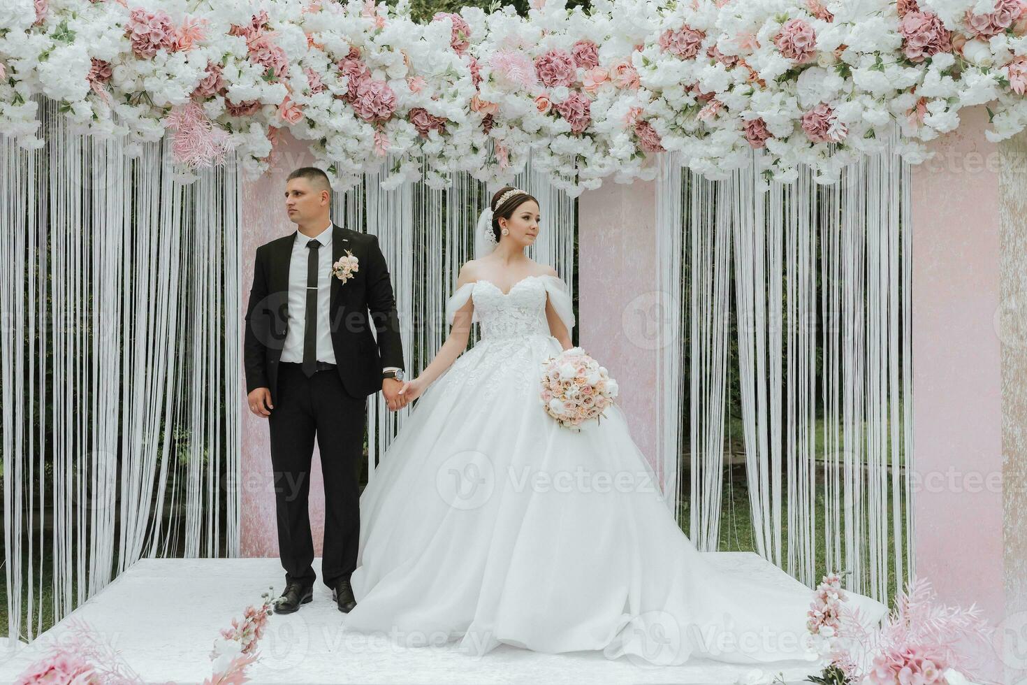 aantrekkelijk bruid en bruidegom Bij de ceremonie Aan hun bruiloft dag met een boog gemaakt van roze en wit bloemen. mooi pasgetrouwden, een jong vrouw in een wit jurk met een lang trein, mannen in een zwart pak. foto