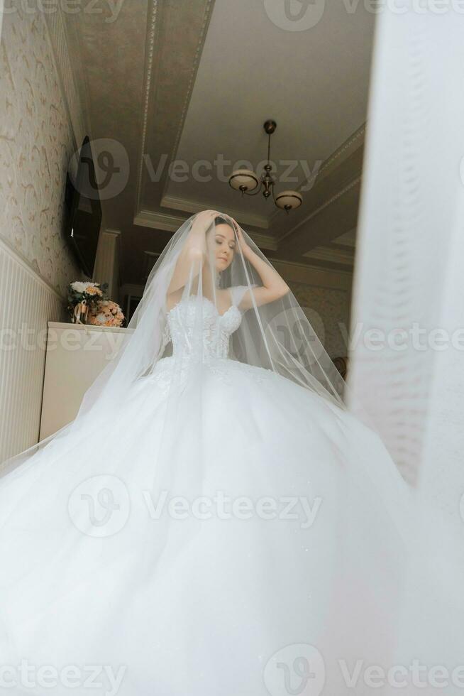 voorbereiding voor de bruiloft. mooi jong bruid in wit luxueus bruiloft jurk, tiara Aan hoofd, lang sluier in Koninklijk hotel kamer. luxe glimlachen model- foto