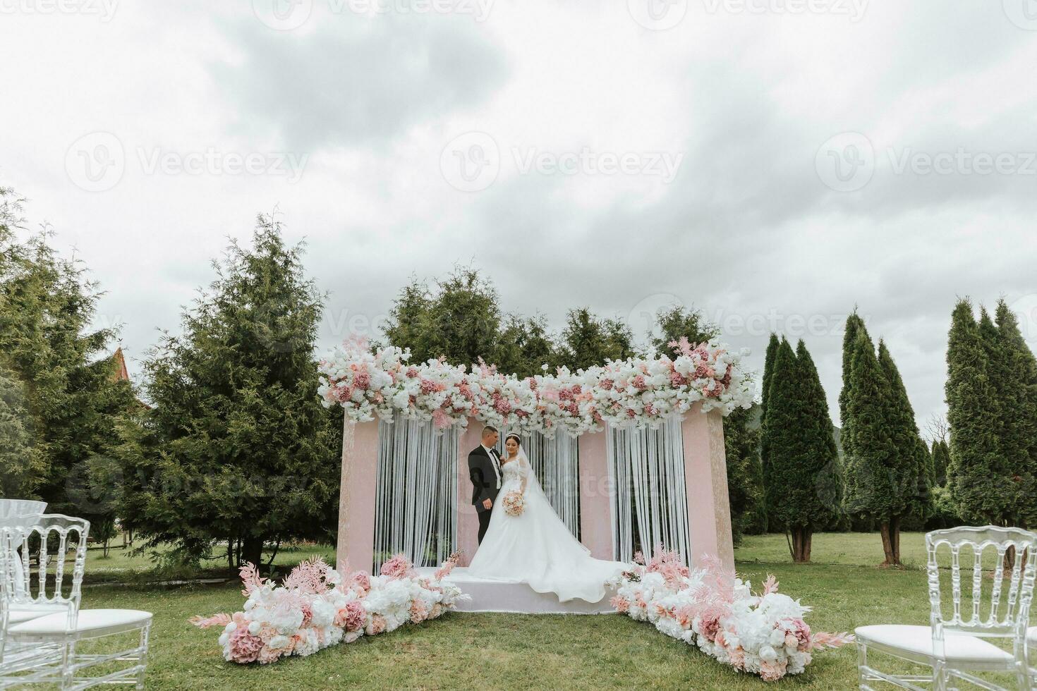 aantrekkelijk bruid en bruidegom Bij de ceremonie Aan hun bruiloft dag met een boog gemaakt van roze en wit bloemen. mooi pasgetrouwden, een jong vrouw in een wit jurk met een lang trein, mannen in een zwart pak. foto