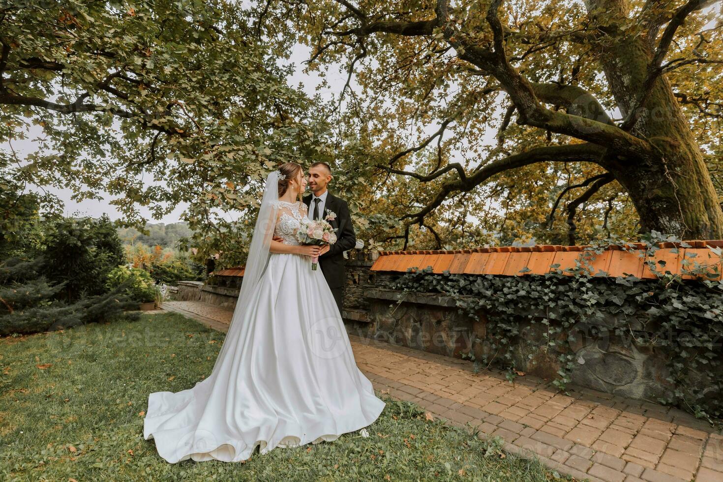 modieus bruidegom en schattig brunette bruid in wit jurk met professioneel bedenken en boeket van bloemen zijn knuffelen, lachend in park, tuin, Woud buitenshuis. bruiloft fotografie foto