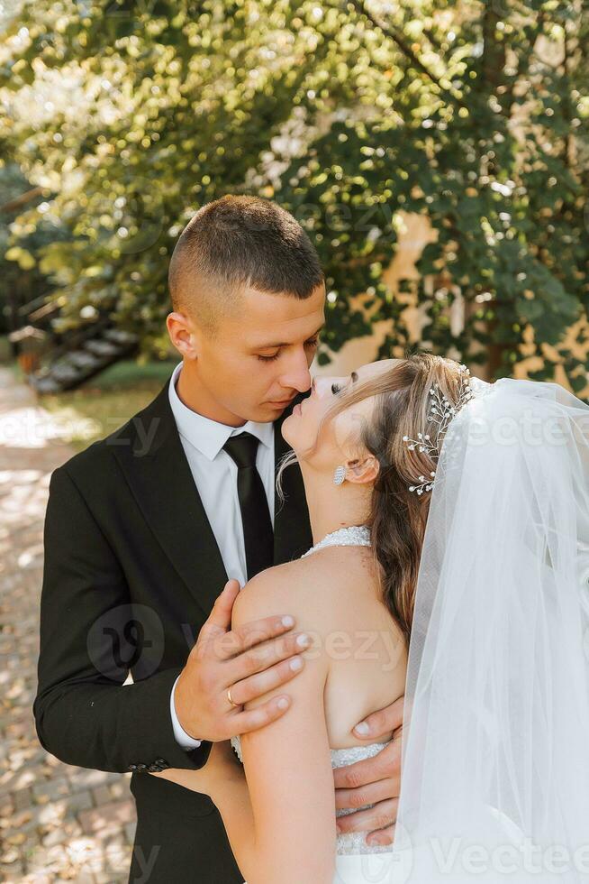 een elegant bruidegom in een zwart pak en een schattig bruid in een wit jurk met een lang sluier zijn knuffelen in een park. bruiloft portret van glimlachen en gelukkig pasgetrouwden. foto