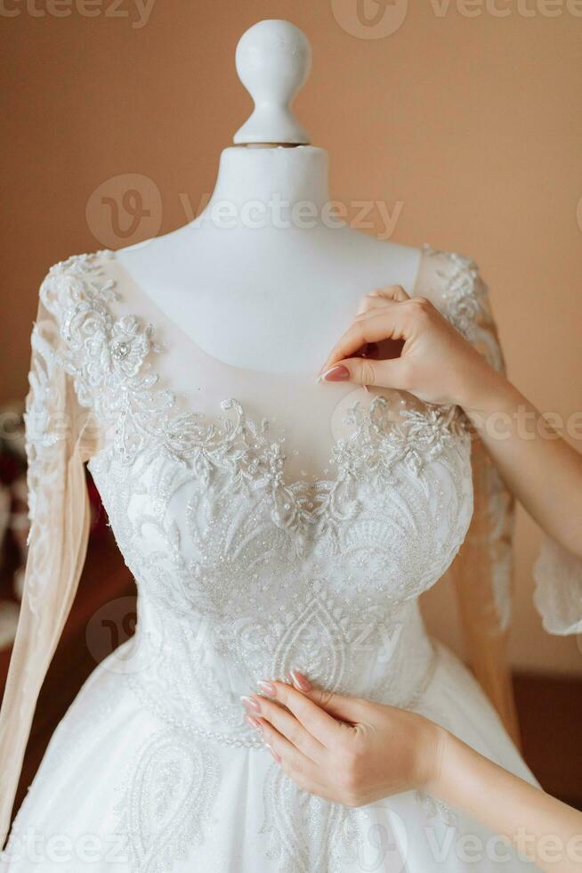 een gelukkig bruid is voorbereidingen treffen voor haar luxueus bruiloft Bij huis, Holding haar mooi jurk in haar handen. portret van een vrouw met professioneel haar- en bedenken en een glimlach in een badjas foto
