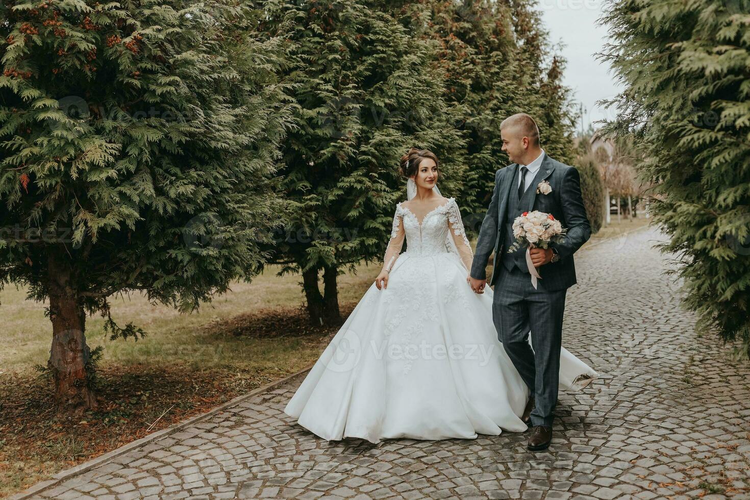 bruidegom en bruid wandelen en Holding handen in de park tussen hoog groen bomen. wijde hoek herfst foto. omvangrijk bruiloft jurk. klassiek pak van de bruidegom foto