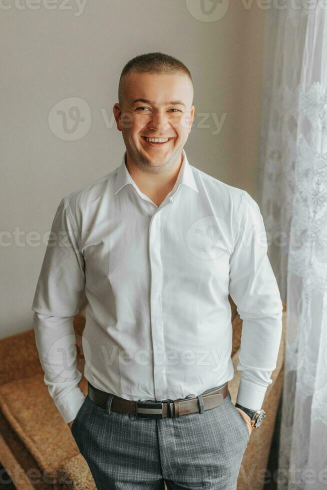 portret van een jong glimlachen Mens in een overhemd in zijn kamer. de bruidegom krijgt gekleed en bereidt zich voor voor de bruiloft ceremonie foto