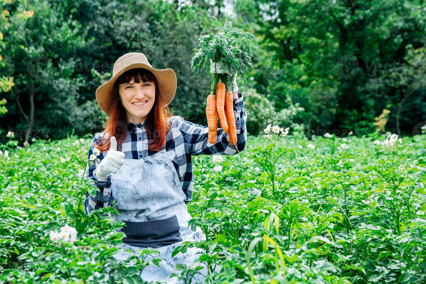 vrouw boer houdt een bos wortelen op de achtergrond moestuin stro hoed en omringd door de vele planten in haar moestuin foto