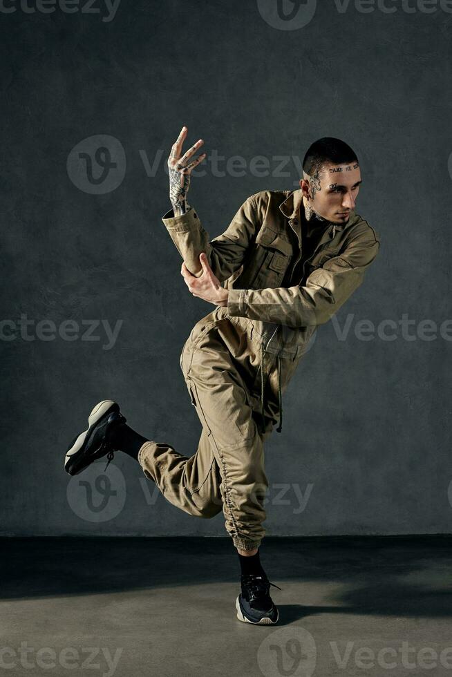 modern Mens met getatoeëerd lichaam en gezicht, oorbellen, baard. gekleed in khaki overall en zwart sportschoenen. dansen Aan grijs achtergrond. danszaal, hiphop foto