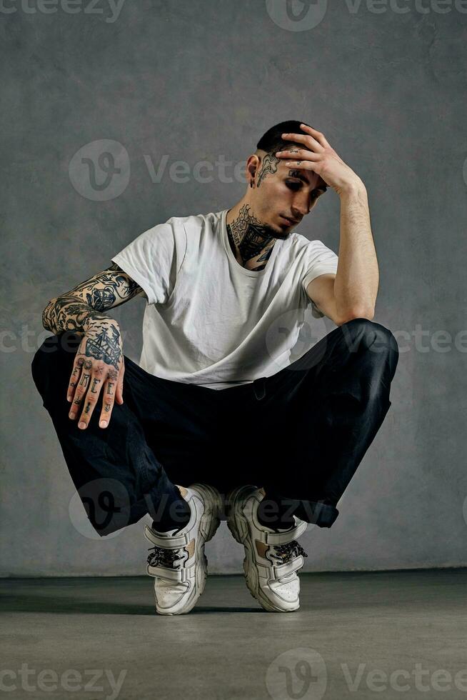 sterk getatoeëerd Mens met baard. gekleed in wit t-shirt, sportschoenen, zwart sport- broek. gehurkt Aan sokken, grijs achtergrond. danszaal, hiphop foto