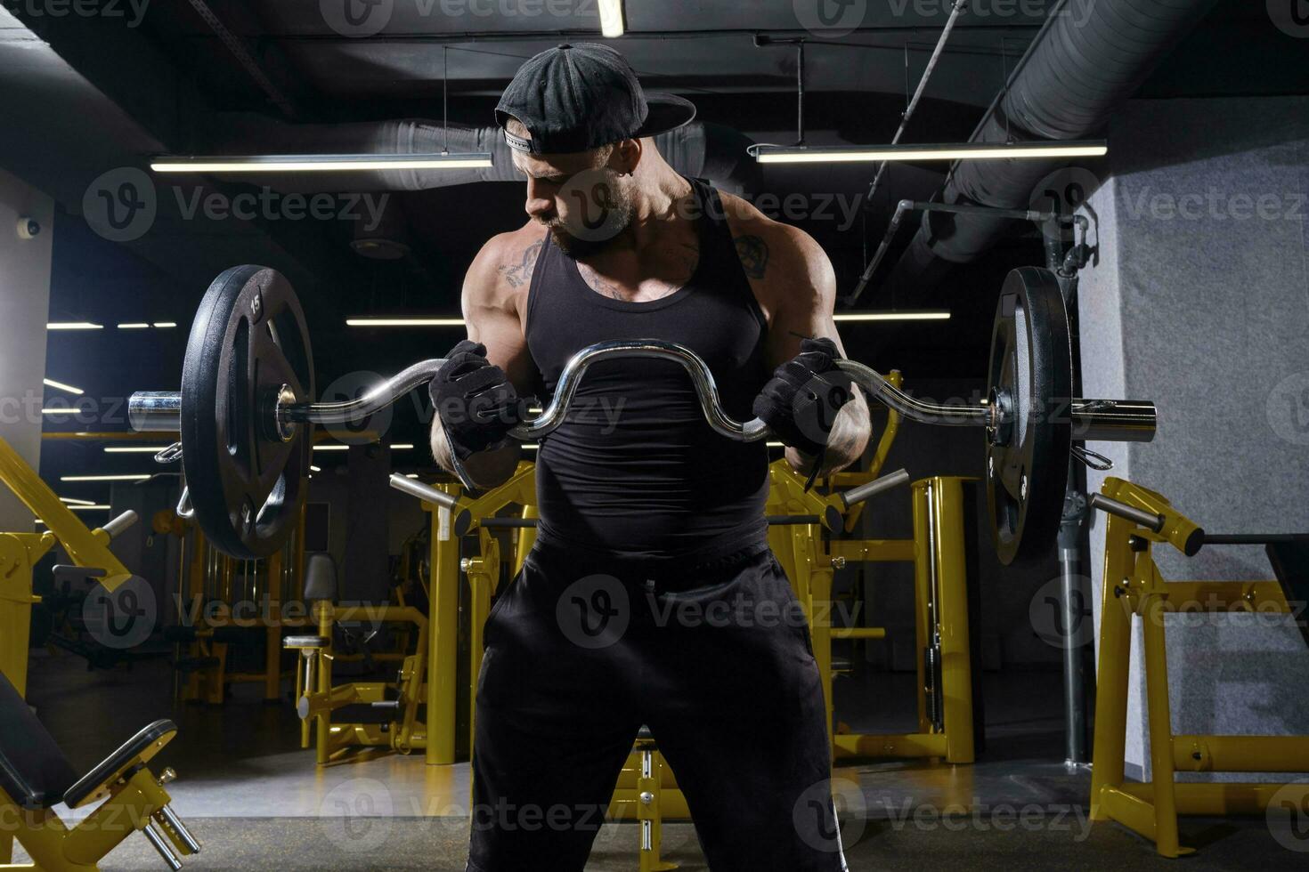 getatoeëerd mannetje in zwart sport handschoenen, korte broek, hesje en kap. hijs- halter, opleiding zijn biceps, staand in donker Sportschool met geel apparatuur. dichtbij omhoog foto