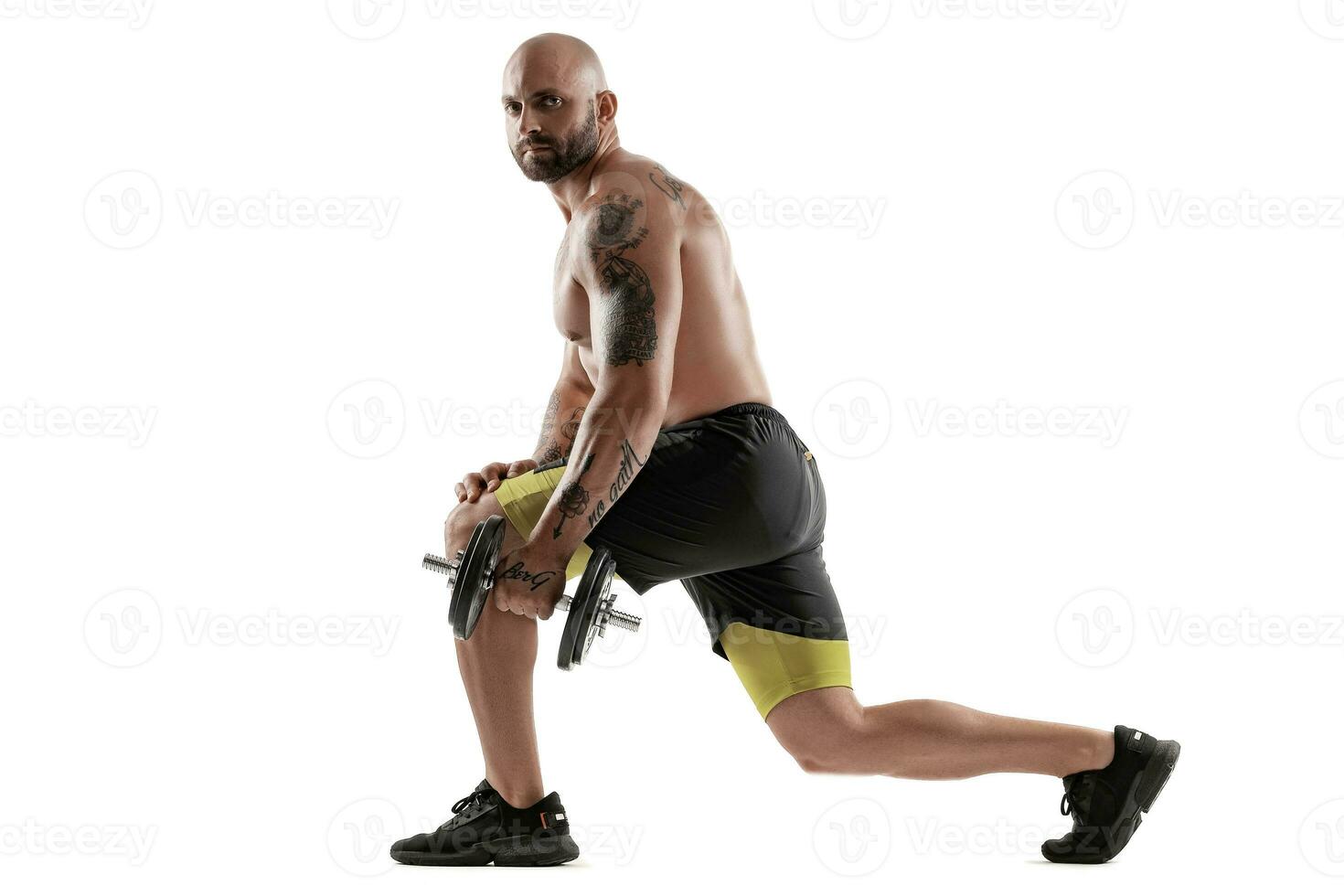 atletisch kaal, getatoeëerd Mens in zwart shorts en sportschoenen is poseren met een halter geïsoleerd Aan wit achtergrond. vol lengte portret. foto