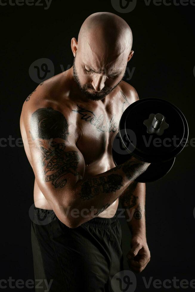 atletisch kaal, baard, getatoeëerd Mens in zwart shorts is poseren met een halter tegen een zwart achtergrond. detailopname portret. foto