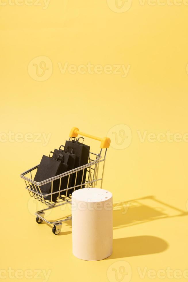 leeg mock podium of voetstuk en miniatuur supermarktkar met zwarte boodschappentassen in black friday-uitverkoop op gele achtergrond foto