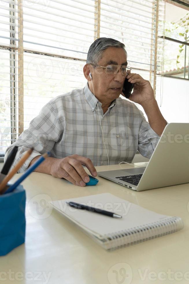 de volwassen man zit thuis aan tafel en gebruikt zijn mobiele telefoon. voor hem staat een laptop foto