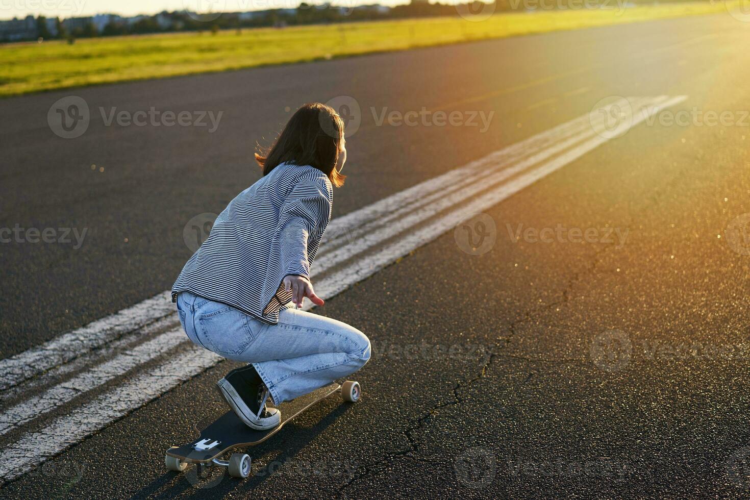kant visie van mooi Aziatisch meisje Aan skateboard, rijden haar kruiser naar de zon Aan een leeg weg. gelukkig jong schaatser genieten van zonnig dag Aan haar vleet foto