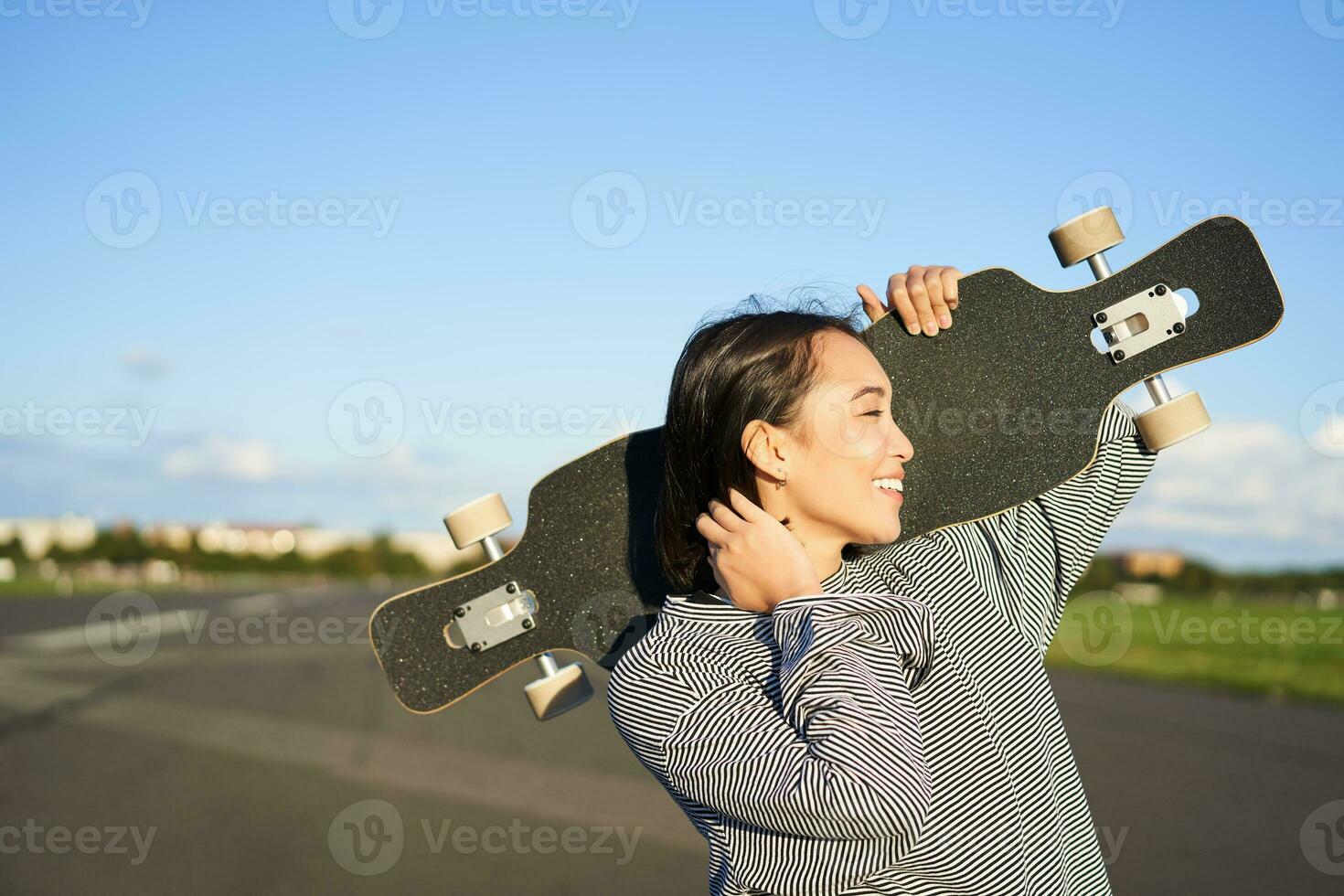 gelukkig en vrij Aziatisch meisje Holding kruiser bord Aan schouders en wandelen naar camera Aan leeg weg, het schaatsen Aan longboard en genieten van zonnig weer foto