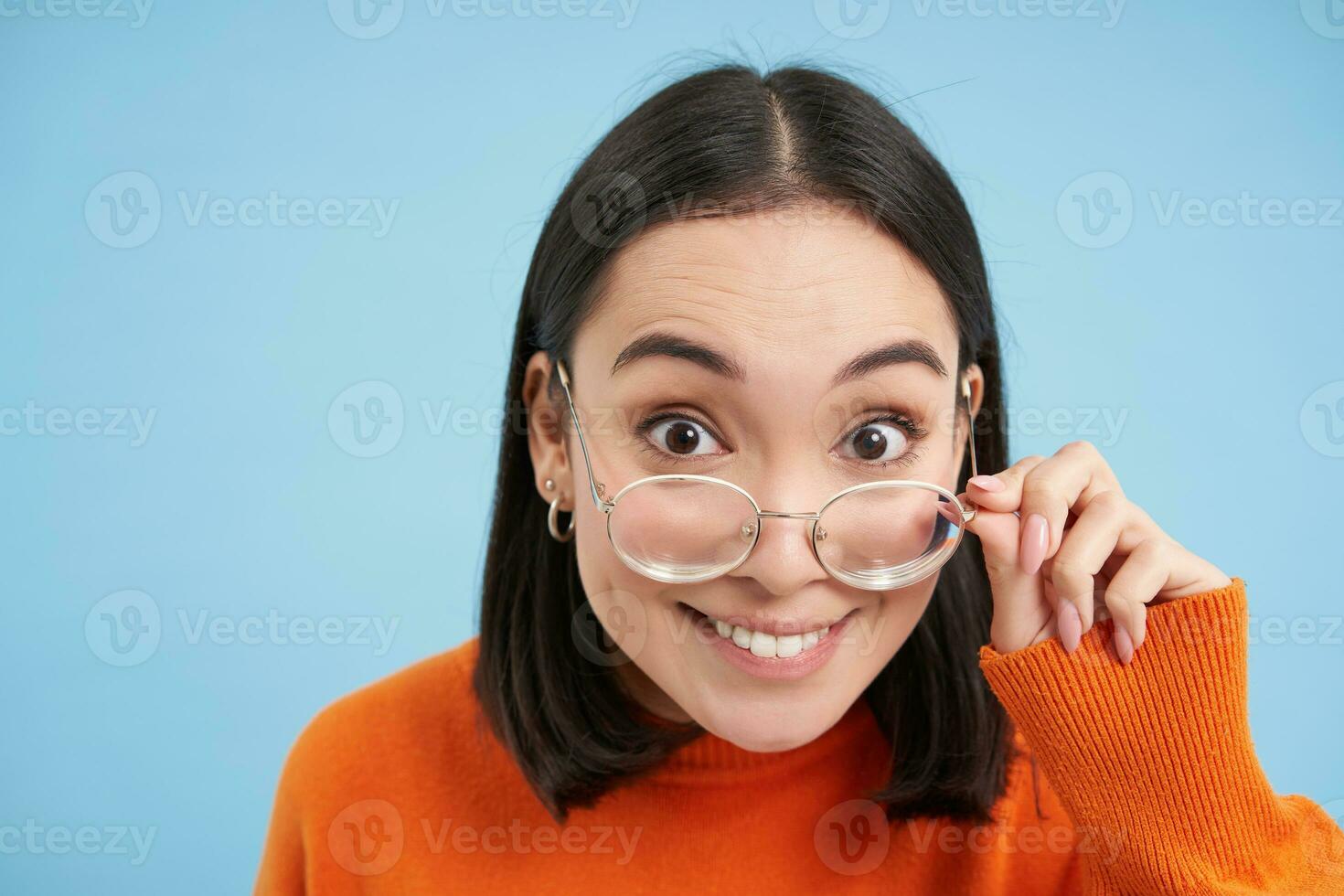 portret van brunette Japans vrouw in bril, glimlachen en op zoek met gelukkig gezicht Bij camera, probeert voorgeschreven brillen, blauw achtergrond foto