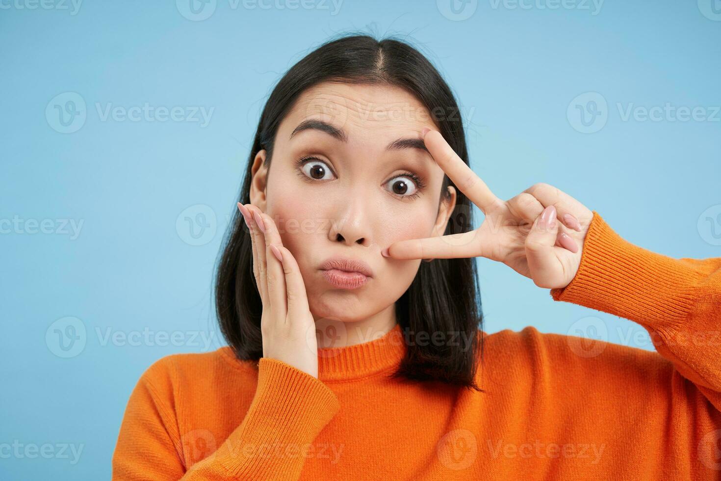 dichtbij omhoog van schattig en dwaas Japans vrouw, shows vrede teken en pruilt, staat over- blauw achtergrond foto