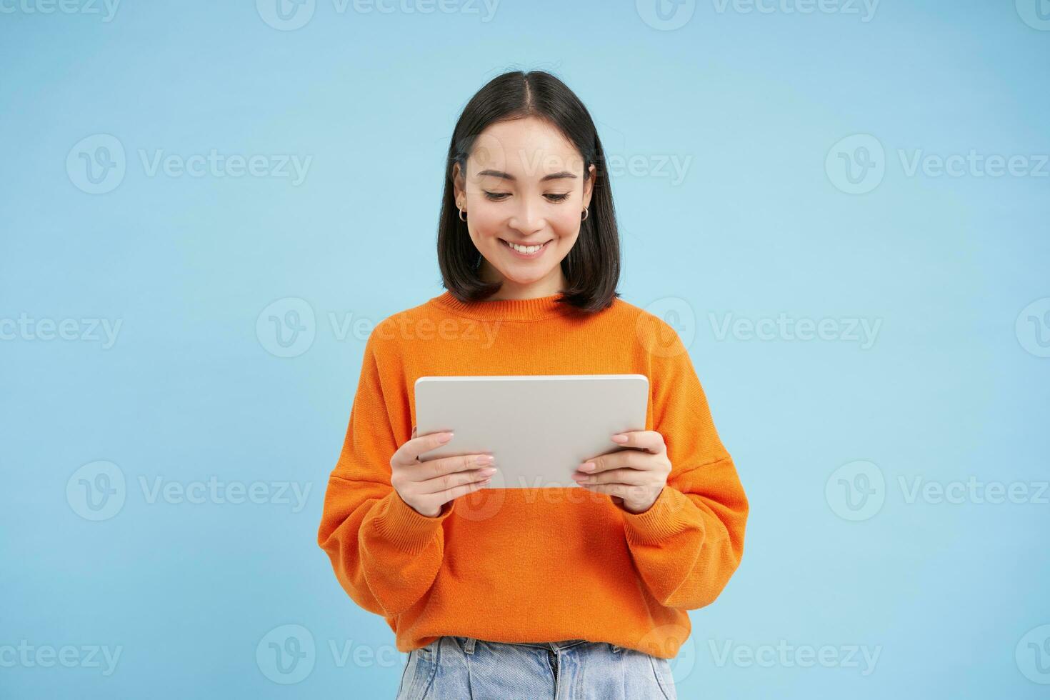 glimlachen mooi Aziatisch vrouw lezing Aan digitaal tablet, aan het kijken videos Aan apparaatje of spelen video spellen, staand over- blauw achtergrond foto
