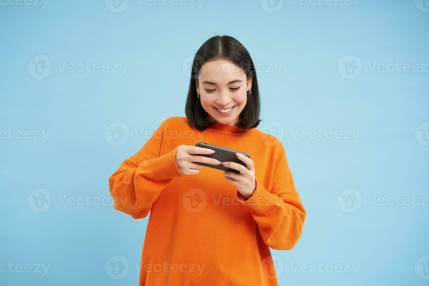 gelukkig glimlachen meisje, 25 jaar, Toneelstukken mobiel video spellen, houdt smartphone in beide handen, staat tegen blauw achtergrond foto