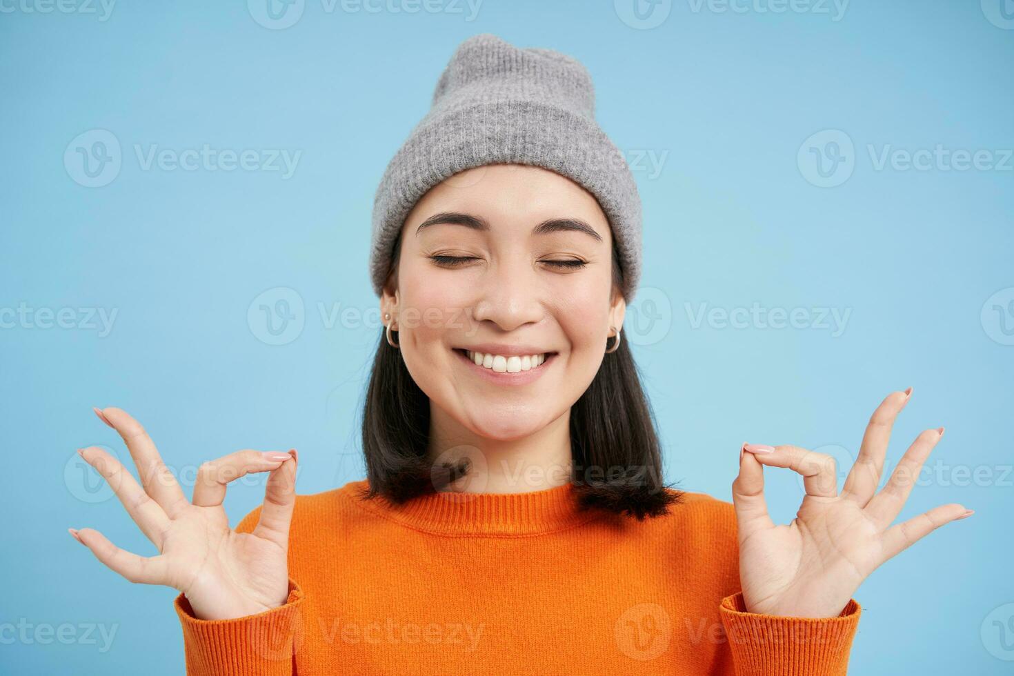 geluiden geest in gezond lichaam. glimlachen kalmte en ontspannen Aziatisch meisje in muts, shows zen, ontspanning gebaar, mediteren, staand over- blauw achtergrond foto