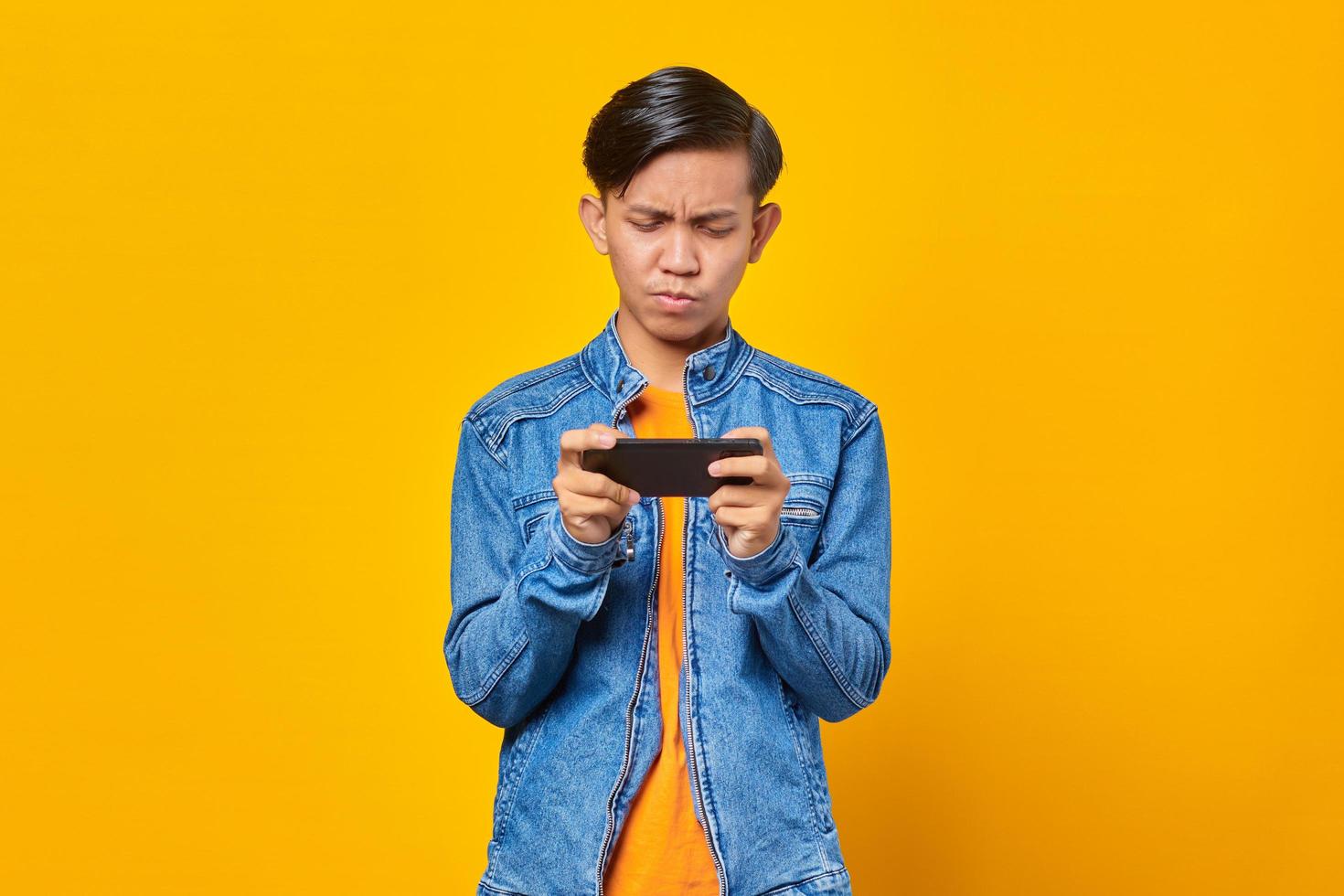 Aziatische man met behulp van mobiele telefoon speelspel met boos gezicht over gele achtergrond foto