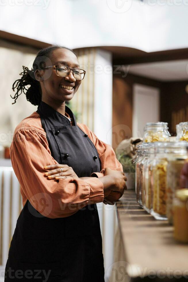 gedetailleerd beeld van een Afrikaanse Amerikaans vrouw winkelier met een schort glimlachen Bij de camera in de buurt glas potten. portret schot van een zwart vrouw vervelend bril met haar arm gekruiste over- haar borst. foto