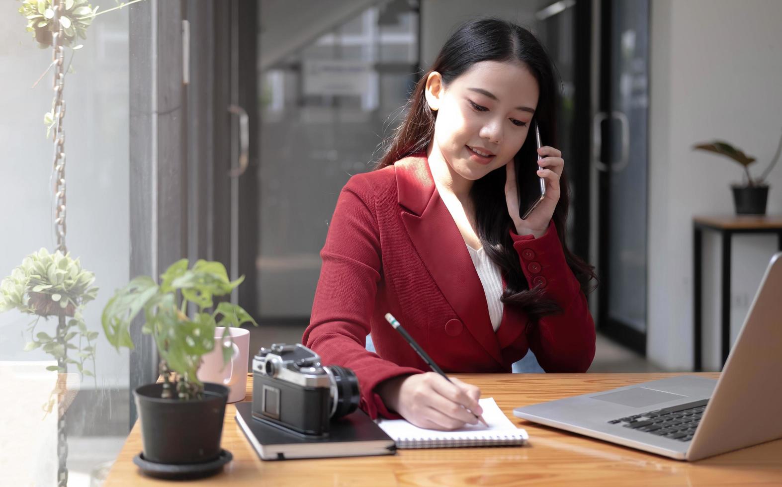 jonge vrouw praten op mobiele telefoon en notities schrijven terwijl ze aan haar bureau zit. aziatische vrouw werkt in kantoor aan huis foto