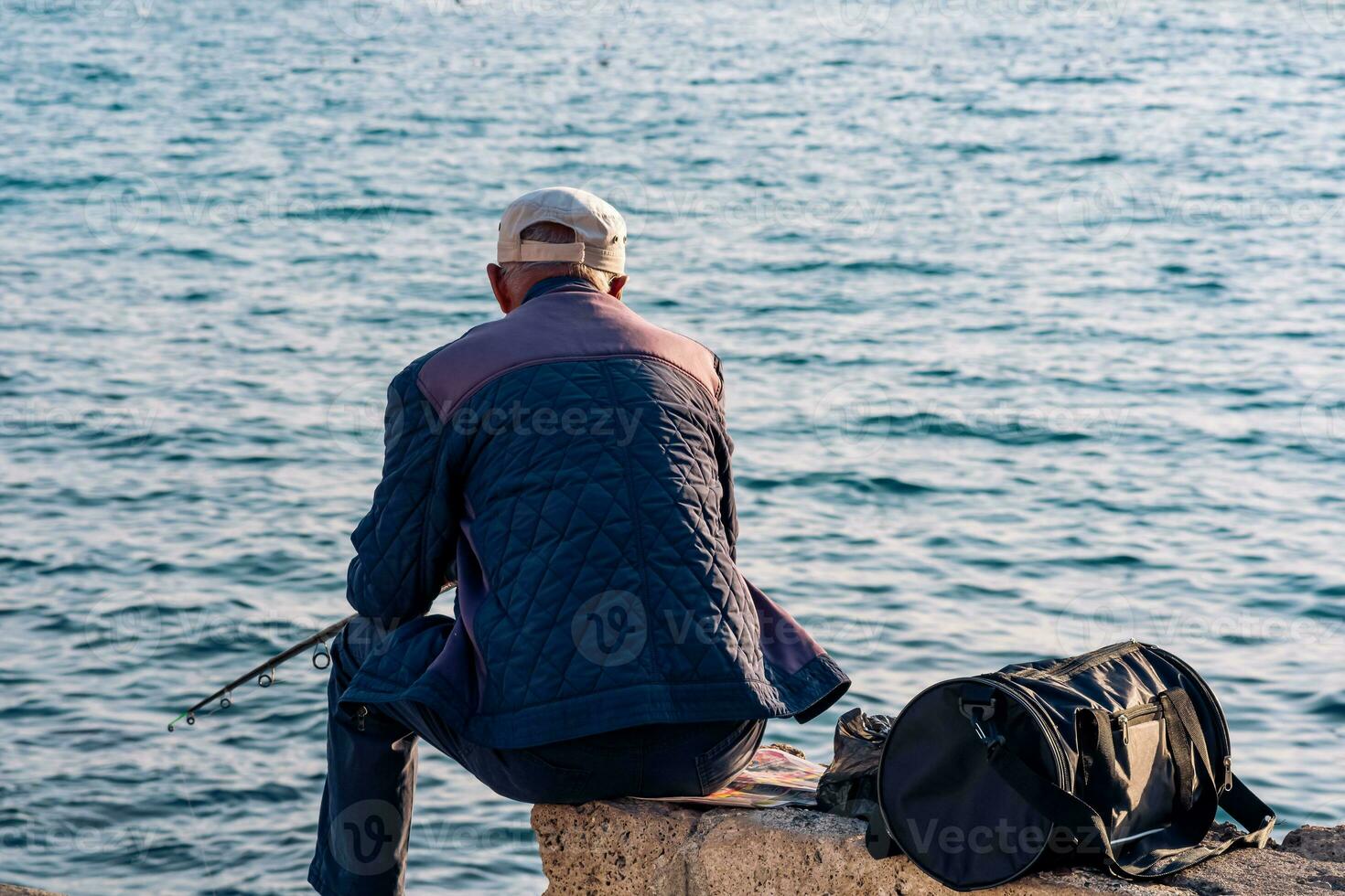 ouderen visser zit met een visvangst hengel Aan een kust- steen foto