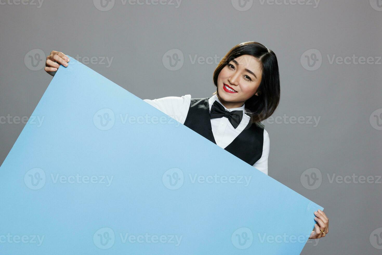 vrolijk Aziatisch vrouw receptioniste in uniform poseren met blanco blauw banier mockup en op zoek Bij camera. glimlachen serveerster Holding leeg whiteboard met copyspace portret foto