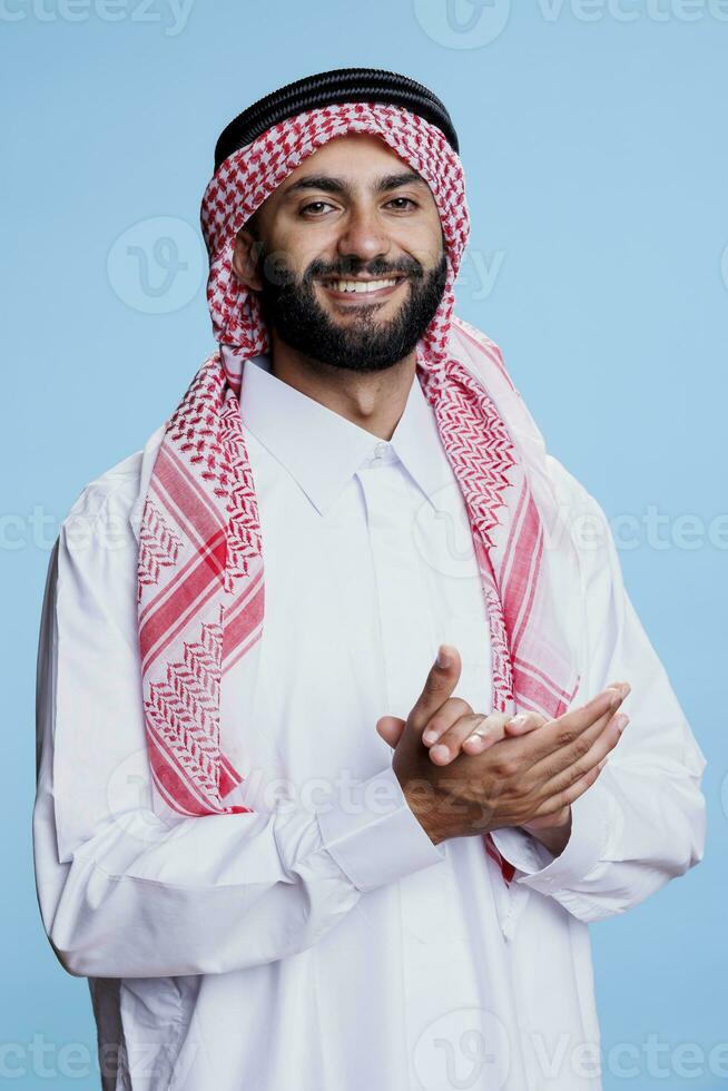 moslim Mens vervelend traditioneel kleren klappen handen en presentatie van geslaagd uitdrukking studio portret. vrolijk Arabisch gekleed in Islamitisch gewaad en hoofddoek applaudisseren en op zoek Bij camera foto