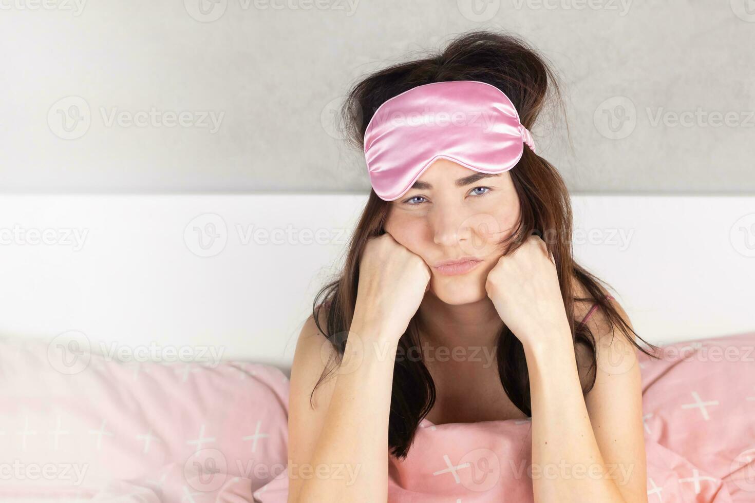 een mooi meisje met blauw ogen zit in bed in pyjama en een slaap masker. vrouw kan niet slaap, slapeloosheid, zorgen, is zenuwachtig, denkt na, heeft nachtmerries roze bed linnen foto