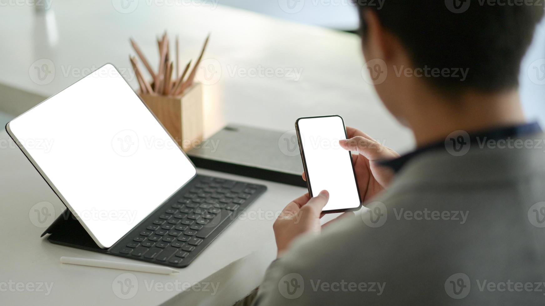 jonge zakenman houdt een smartphone met een leeg scherm in de hand en een laptop met een leeg scherm op het bureau. foto