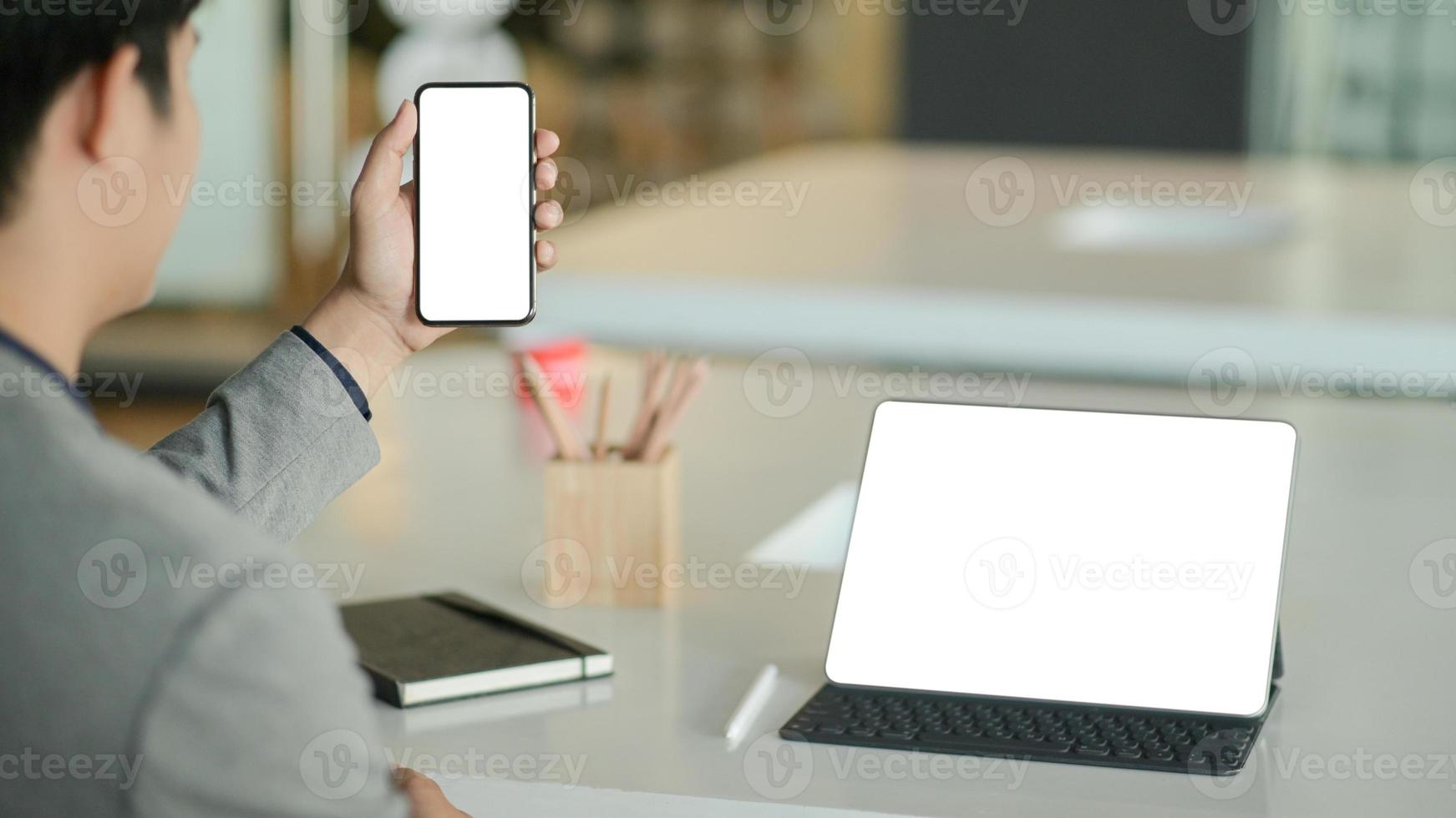 jonge zakenman houdt een smartphone met een leeg scherm in de hand en een laptop met een leeg scherm op het bureau. foto