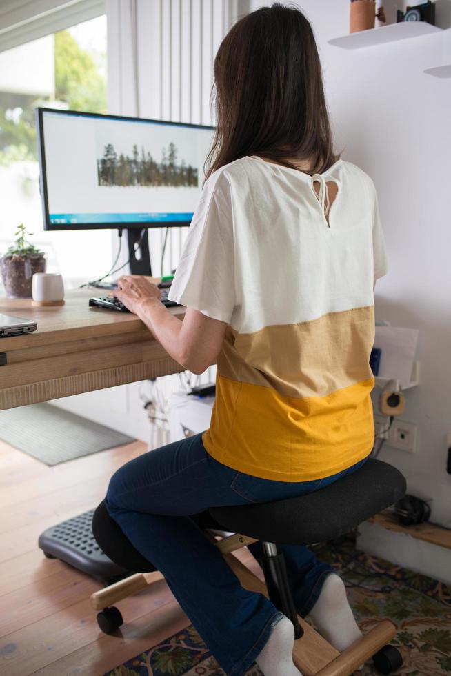 jonge vrouw die thuis werkt met behulp van een ergonomische knielende stoel, gezien vanaf haar rug. foto