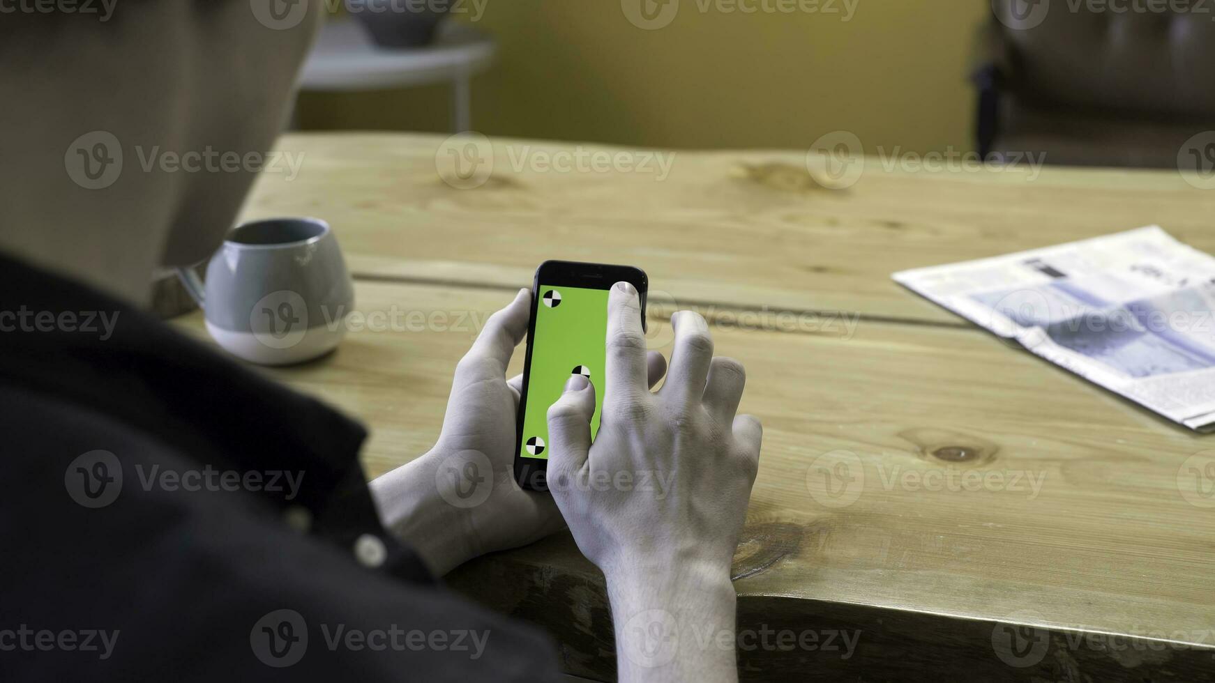 een vent Holding in armen elektronica apparaatje met groen scherm Chroma sleutel, scrollen en tikken het Aan houten tafel achtergrond. voorraad filmmateriaal. menselijk zitten en gebruik de handheld mobiele telefoon in kamer. foto