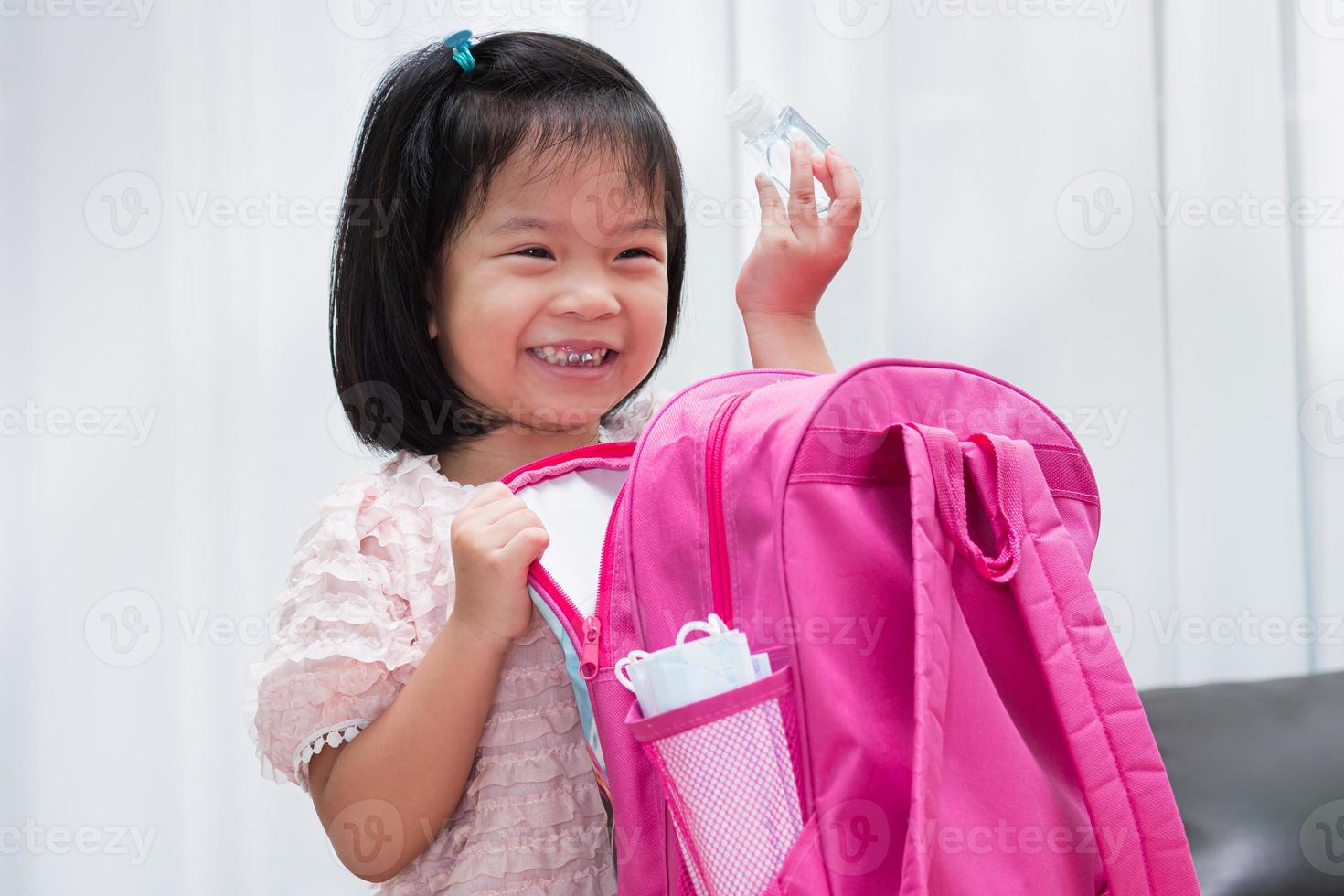 een schattig meisje heeft plezier met het inpakken van haar koffers naar school. meisje met een fles alcoholgel. het hygiënische masker zit naast de zak. foto