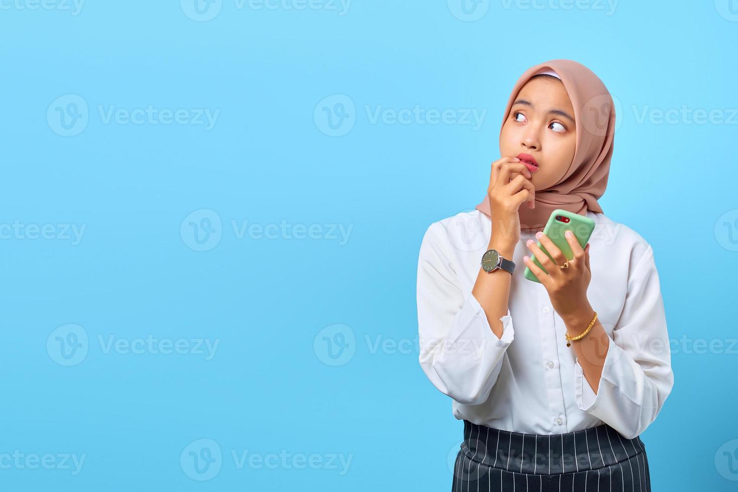 portret van jonge aziatische vrouw die aan iets denkt en mobiele telefoon over blauwe achtergrond houdt foto