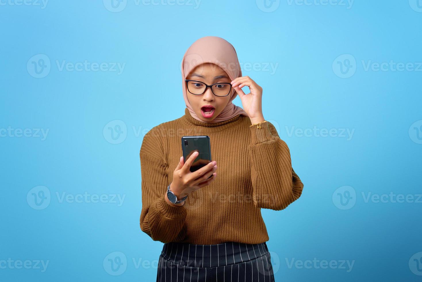 verraste jonge aziatische vrouw die naar het scherm van de mobiele telefoon kijkt met open mond op blauwe achtergrond foto