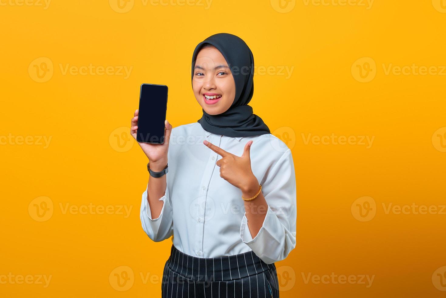 portret van vrolijke jonge aziatische vrouw die het lege scherm van de smartphone op gele achtergrond toont foto