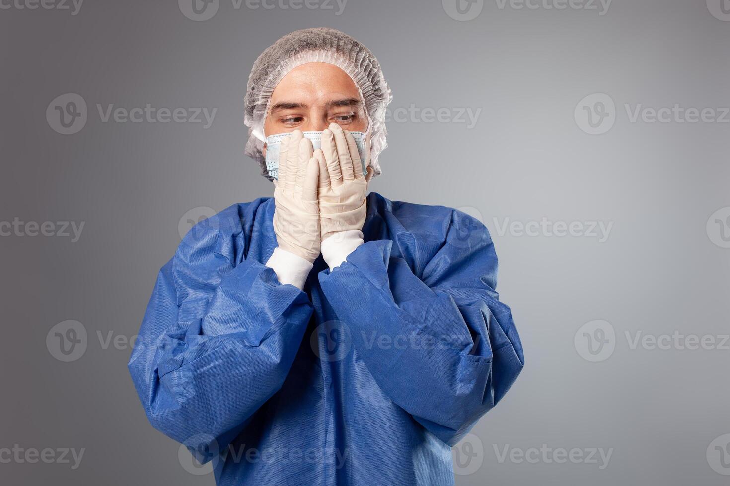 een knap mannetje chirurg is een dokter Aan een grijs achtergrond in de studio. een Mens in een gewaad, een medisch masker, een hoed. leeg ruimte. een Mens covers zijn mond met zijn handen, doet niet willen naar zeggen iets. foto