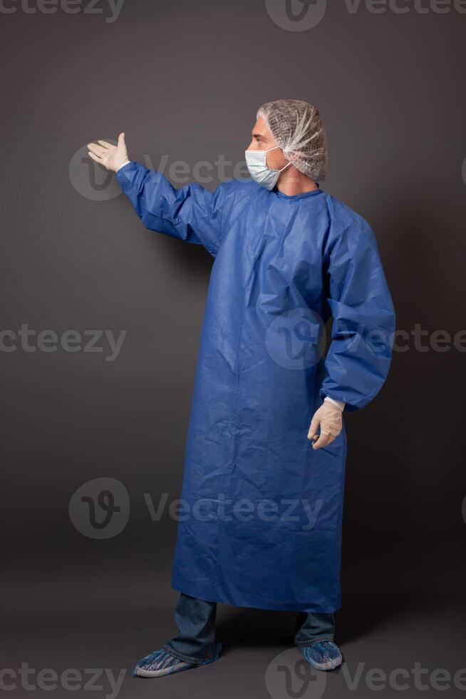 de dokter shows Aan iets met zijn hand- Aan een grijs achtergrond, een leeg ruimte voor tekst. een Mens in een blauw gewaad, hoed, handschoenen, bahillah, in een medisch masker. foto