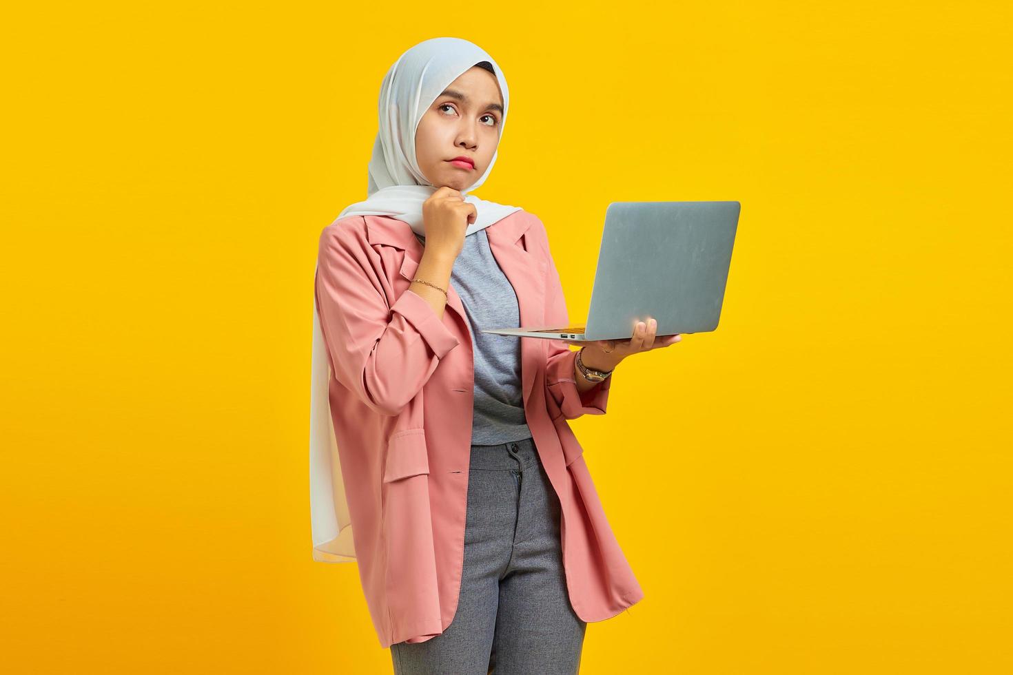 portret van mooie jonge aziatische vrouw met laptop en peinzende kin geïsoleerd over gele achtergrond foto