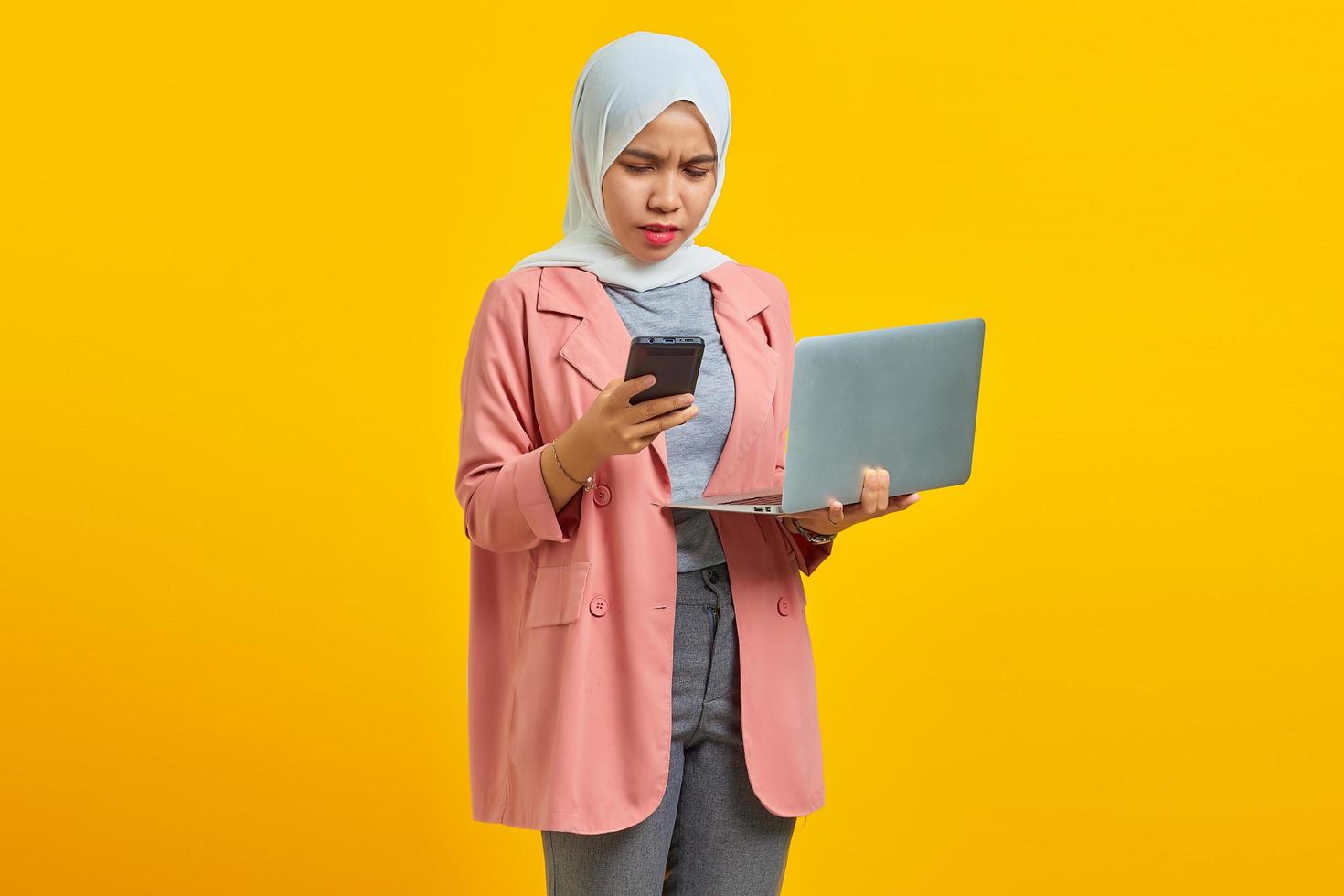boze jonge vrouw die laptop en mobiele telefoon gebruikt terwijl ze geïsoleerd op gele achtergrond staat foto