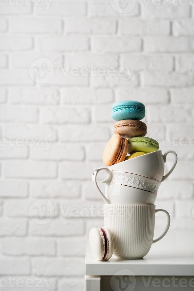 stapel witte koffiekopjes gevuld met bitterkoekjes op de rand van de tafel, witte bakstenen muur achtergrond foto