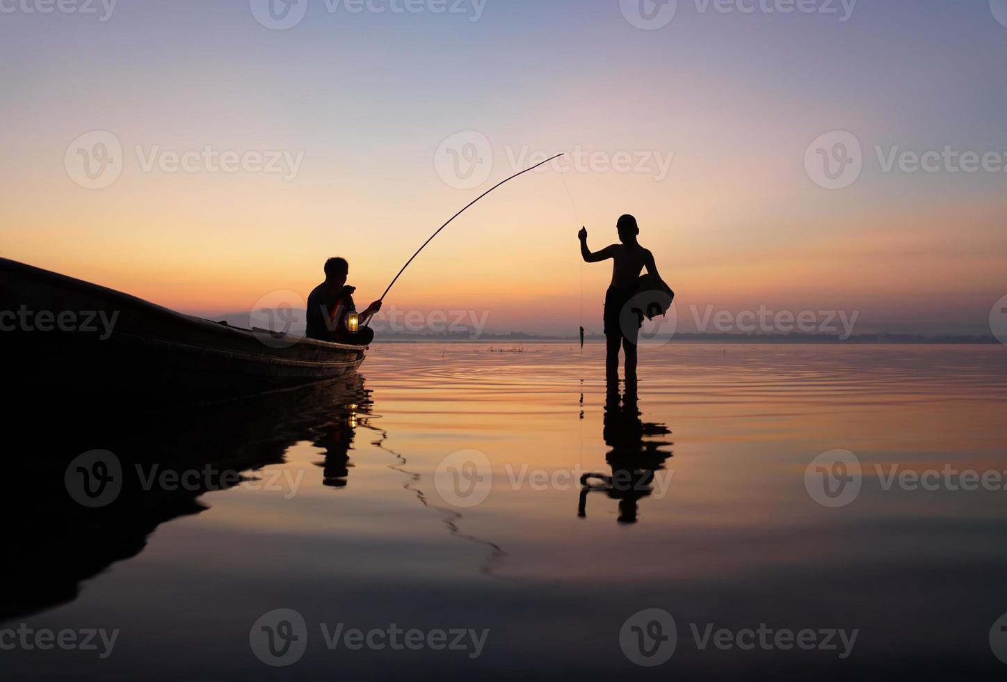 aan de kant van het meer, Aziatische visser zittend op een boot terwijl zijn zoon staat en een hengel gebruikt om vis te vangen bij zonsopgang foto